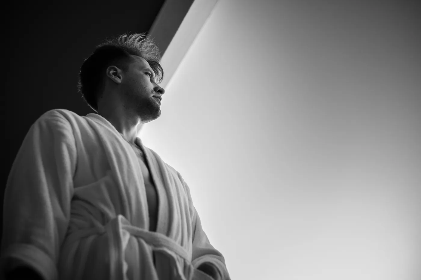 Black-and-white portrait of a man in a Martila Arts Uniform looking upward by a bright window, with a moody, contemplative feel.
