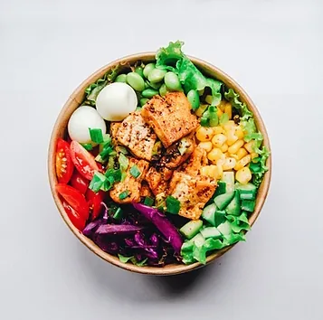 Top view of a colorful poke bowl with grilled tofu, edamame, corn, avocado, tomato, and purple cabbage on a light background.