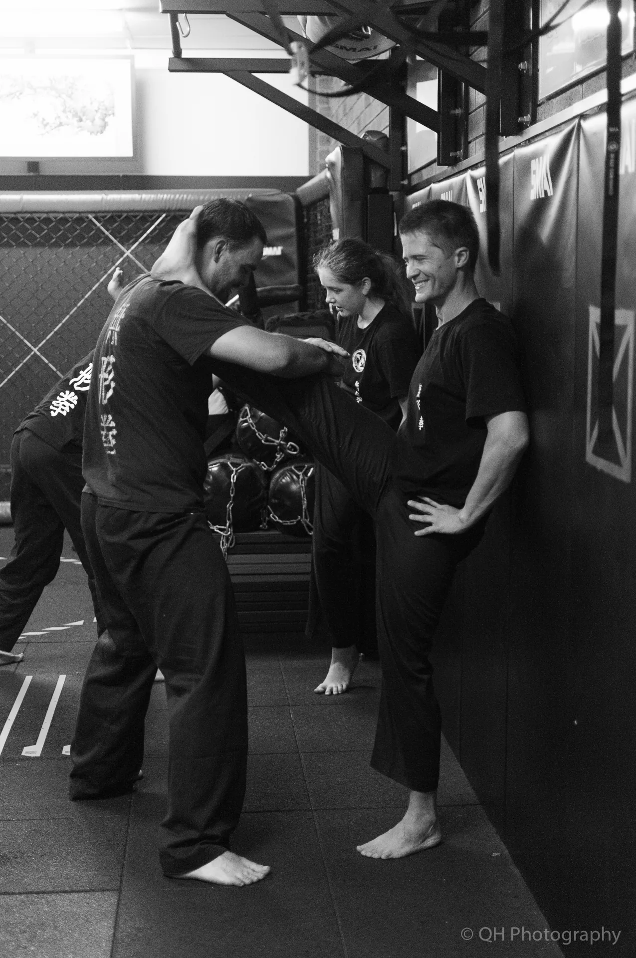 Black-and-white martial arts class, three people stretching barefoot on mats in a gym, smiling by padded wall; QH Photography watermark.