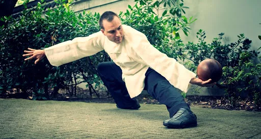 Man in white martial arts uniform crouches low performing a Kung Fu pose, balancing with one hand holding a wooden ball, against green shrubs.
