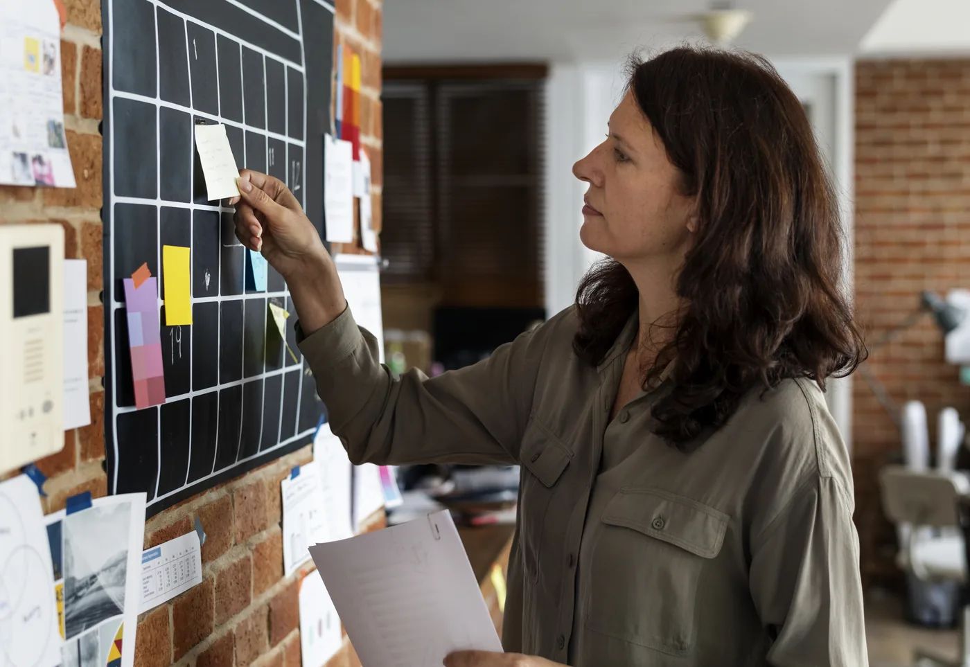 Woman in an office pinning a sticky note to a black grid planning board, holding papers, focused and thoughtful.