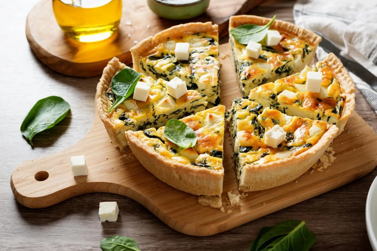 Spinach and cheese quiche slices on a wooden board, garnished with basil and cheese cubes in a warm kitchen scene.