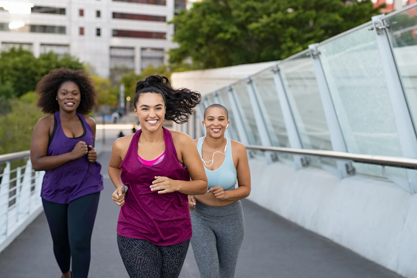 Three women jog smiling on a city bridge, wearing purple, magenta, and blue workout gear, with buildings and trees behind them.