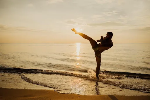Silhouetted man does a high kick on a beach at sunset, with calm waves and golden sky behind him.