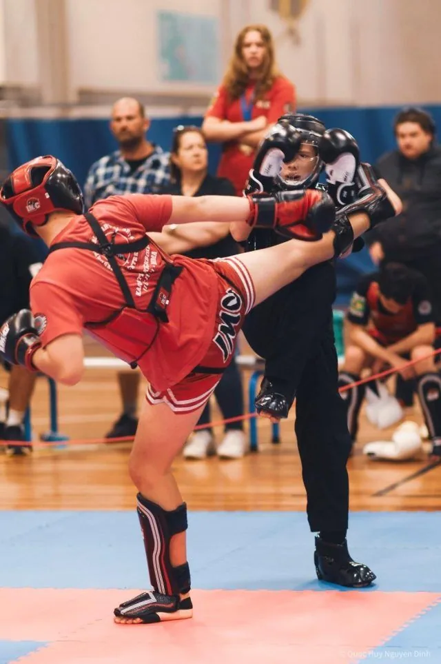 Two kickboxers in red and black gear spar in a gym, one landing a high kick as spectators watch intently.