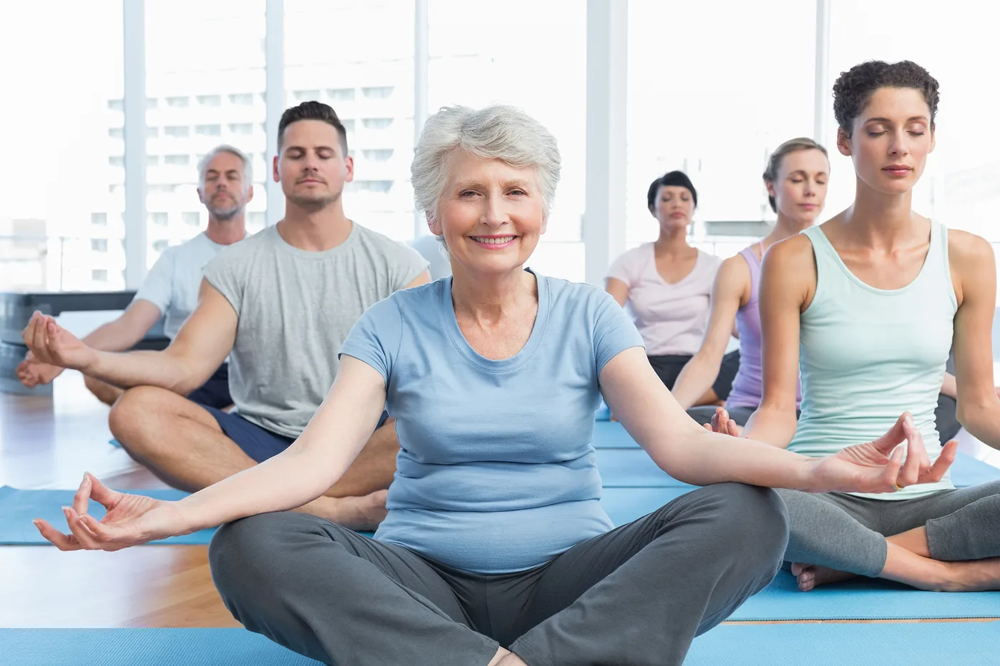Group meditation class in a bright studio, with an older woman smiling at center and others seated cross-legged, eyes closed.