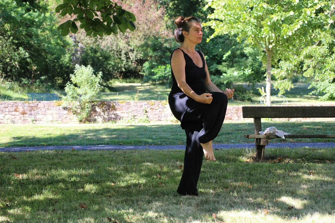 Woman in black Tai Chi clothes balances on one leg in a sunny park beside a bench, surrounded by green trees.