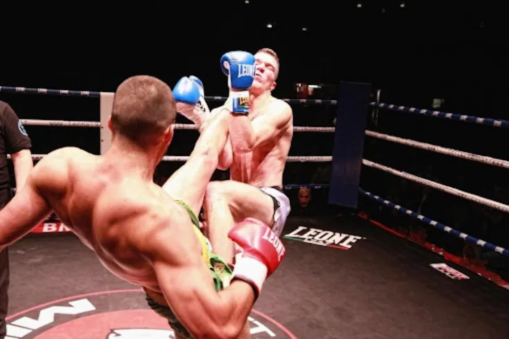 Two kickboxers exchange a flying knee and guard in a ring, with LEONE logos on the ropes and mat under bright lights.