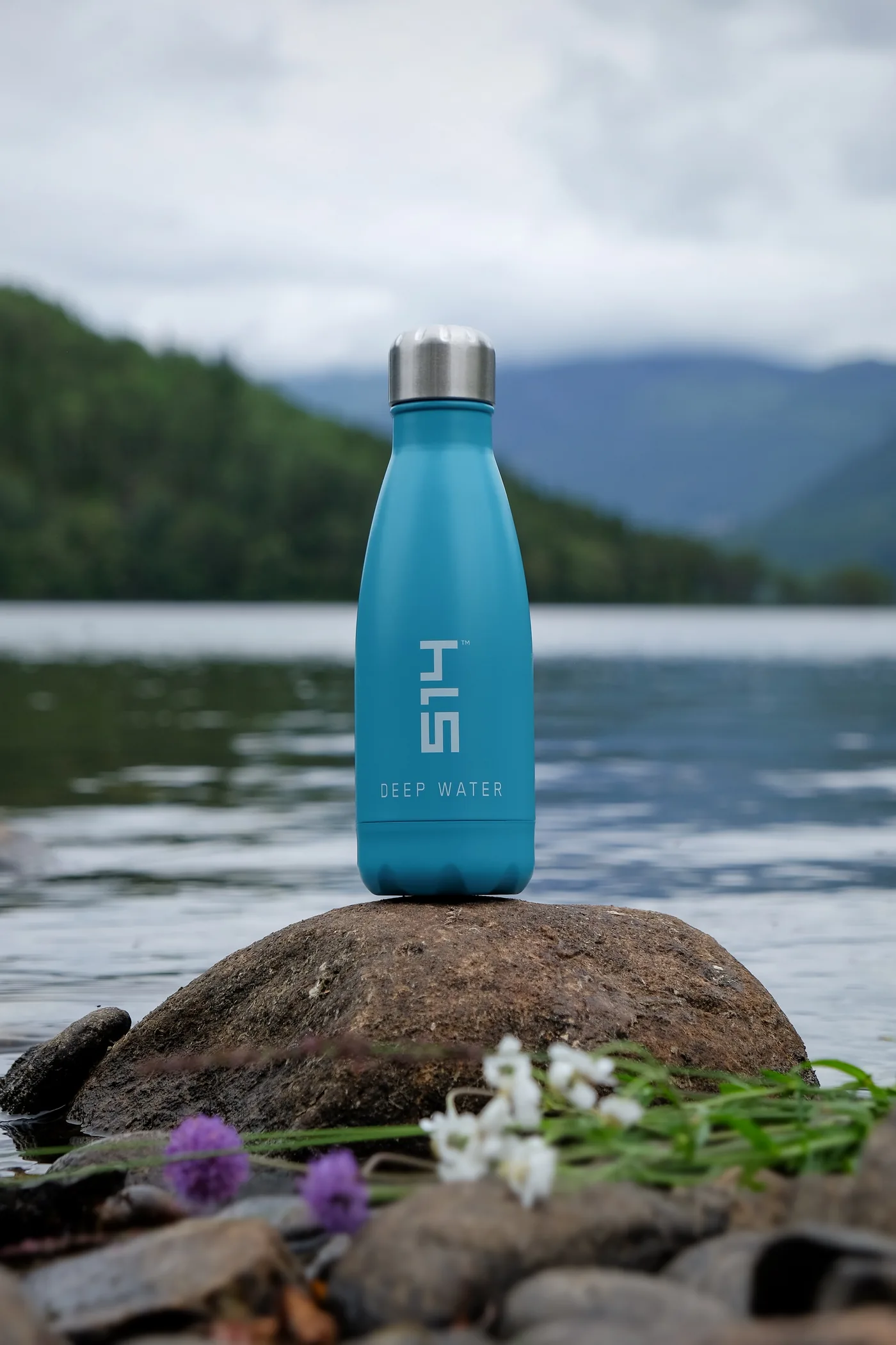 Blue reusable bottle labeled DEEP WATER sits on a rock by a lake, with blurred mountains, water, and wildflowers behind.