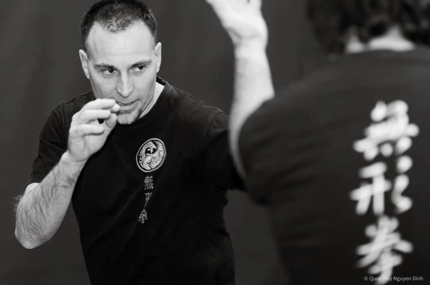 Black-and-white photo of two men practicing martial arts; foreground man in guard pose, with Chinese Kanji characters on black shirts.