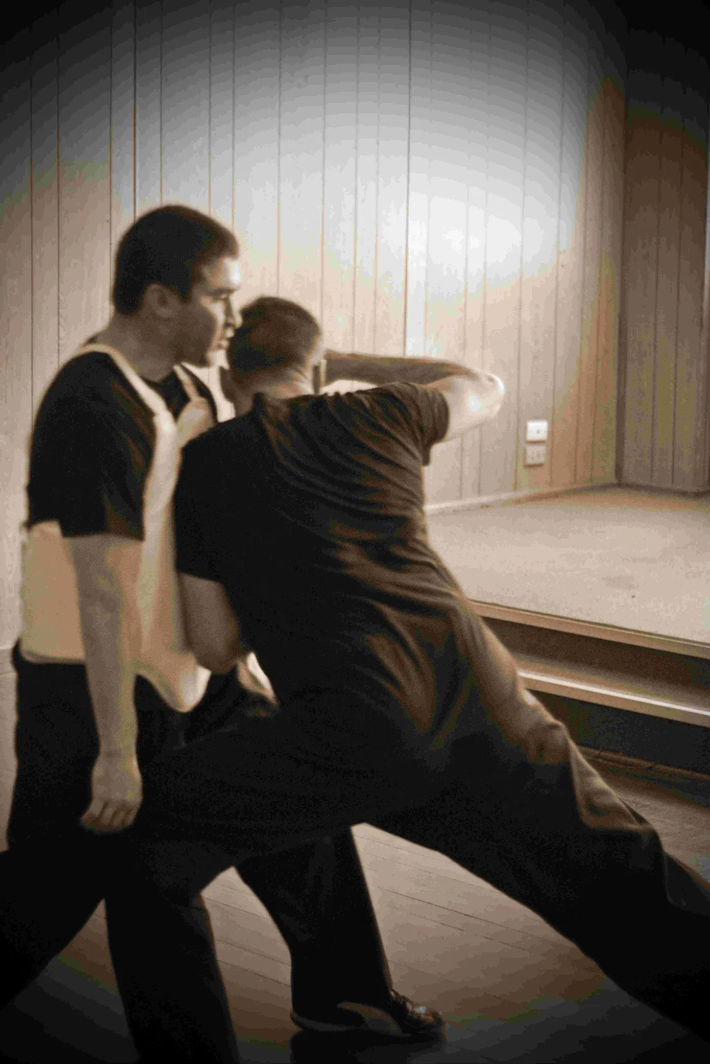 Two men practicing steel body training in a wood-paneled room, one lunging forward to perform a shoulder strike on opponent, under warm light.