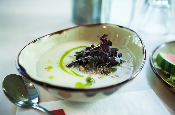 Creamy soup in a bowl topped with dark greens and seeds, green oil drizzle, with a spoon on a bright table setting.