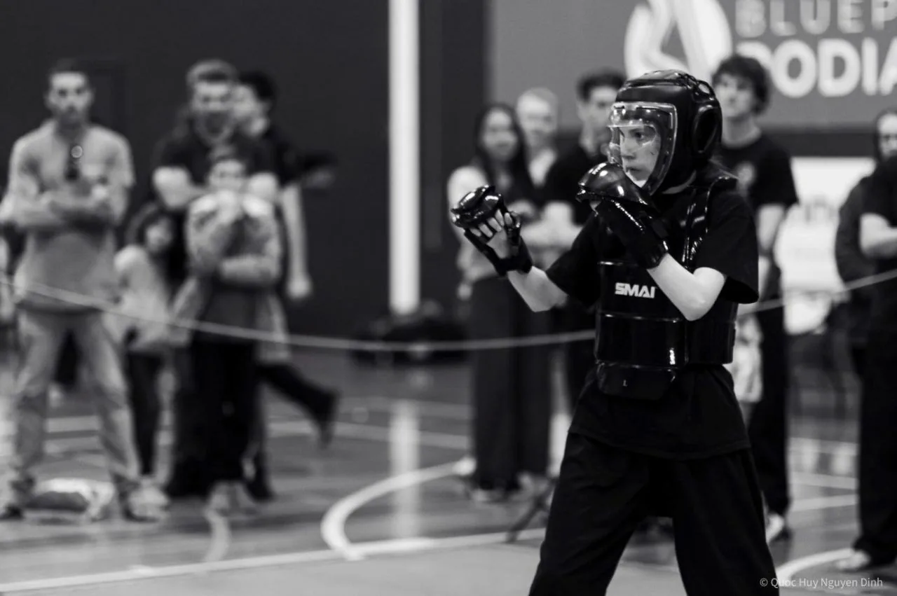 Black-and-white photo of a child in sparring gear and helmet labeled SMAI, hands raised at a Martial Arts tournament with blurred spectators in the background.