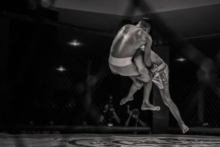 Black-and-white MMA fight in a cage, one fighter lifting and throwing another mid-air, intense action on the mat.