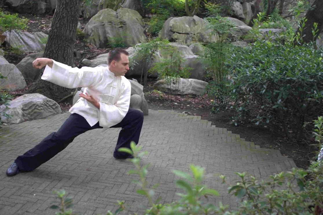Man in a white martial arts outfit holds a low tai chi pose on a stone path in a lush garden with rocks and shrubs.