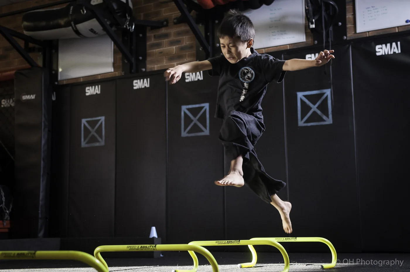 Young boy in black martial arts uniform leaps over yellow hurdles in a gym, with SMAI mats and a focused expression.