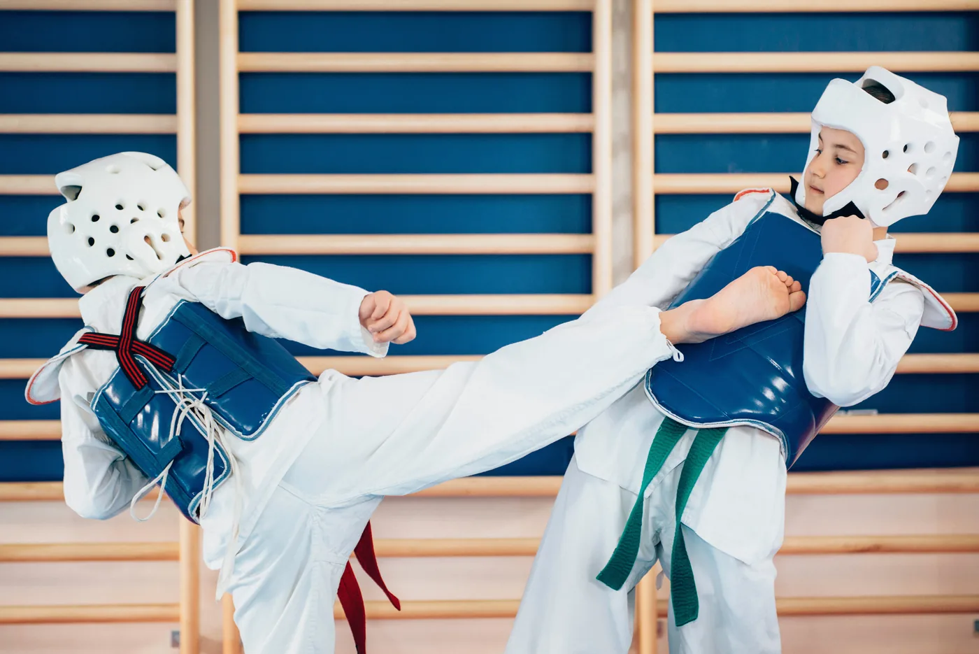 Two children in taekwondo gear sparring indoors, one landing a side kick on the other, with wooden wall bars behind them.