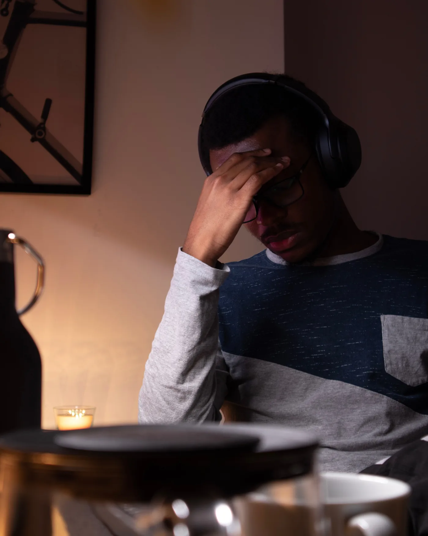 Man wearing headphones and glasses rests his hand on his forehead at a candlelit table, looking tired or stressed.