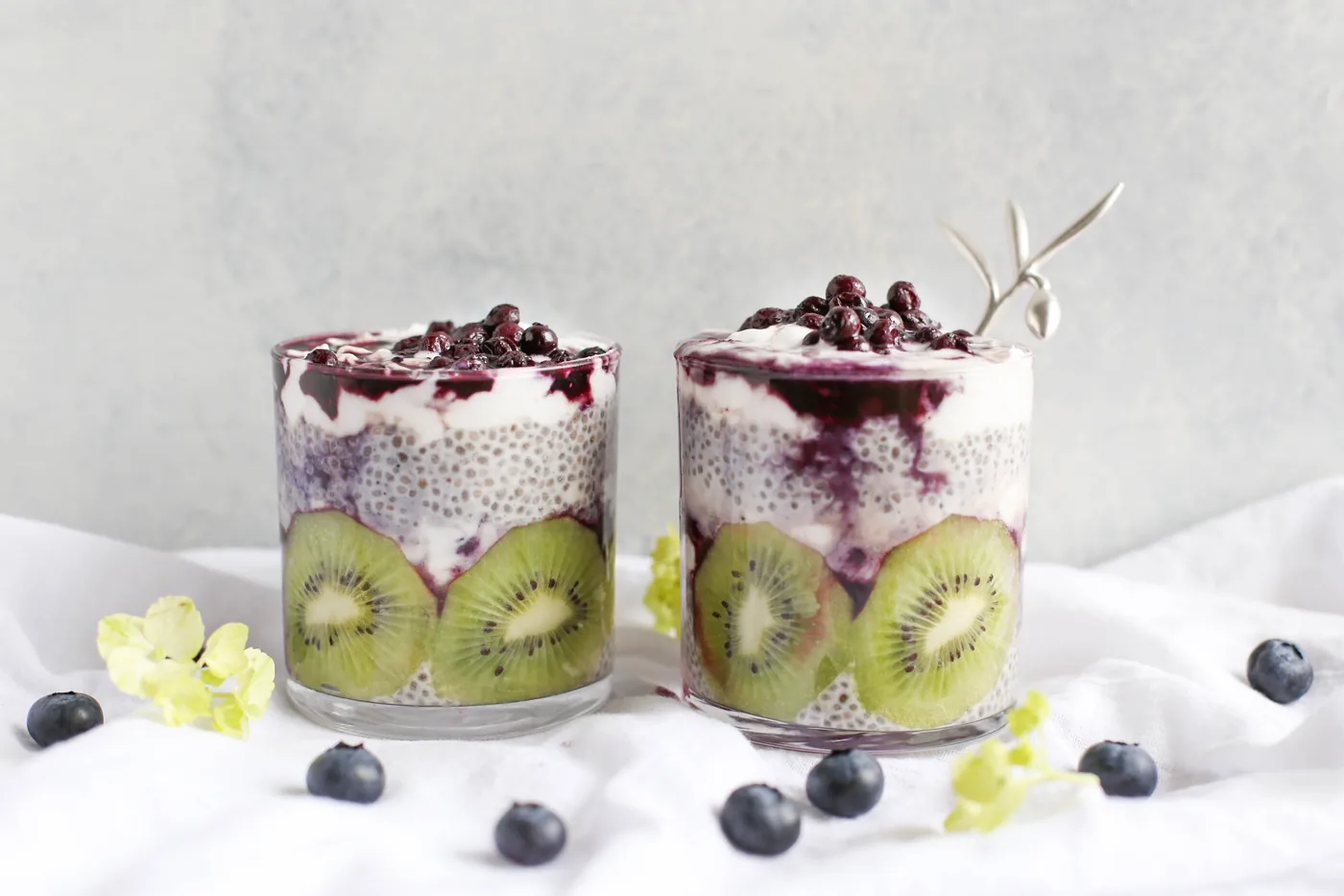 Two glasses of layered chia pudding with kiwi and blueberry sauce on a white cloth, with scattered blueberries and flowers.