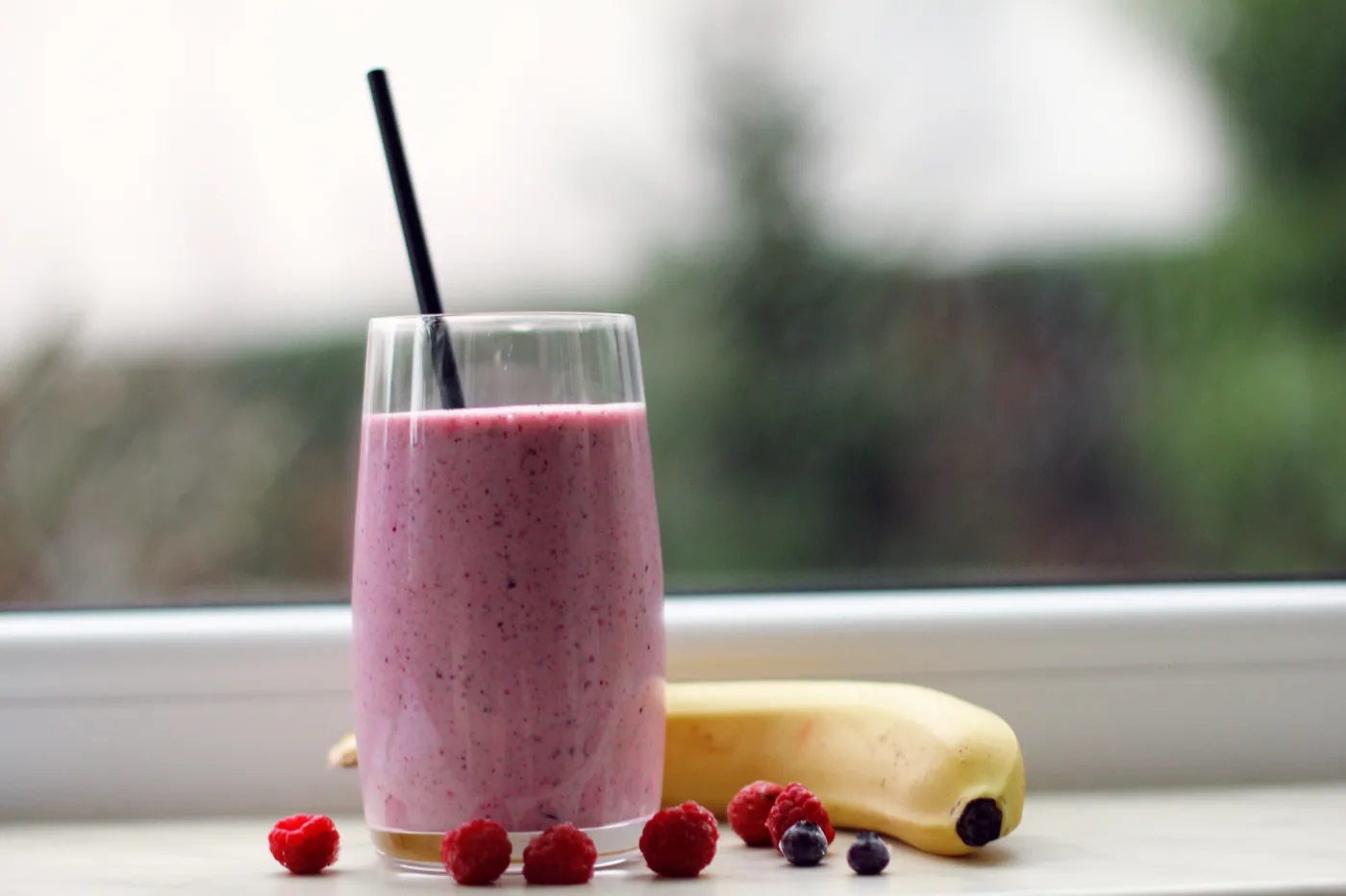 Pink berry smoothie in a glass with a black straw, beside a banana and raspberries on a windowsill.