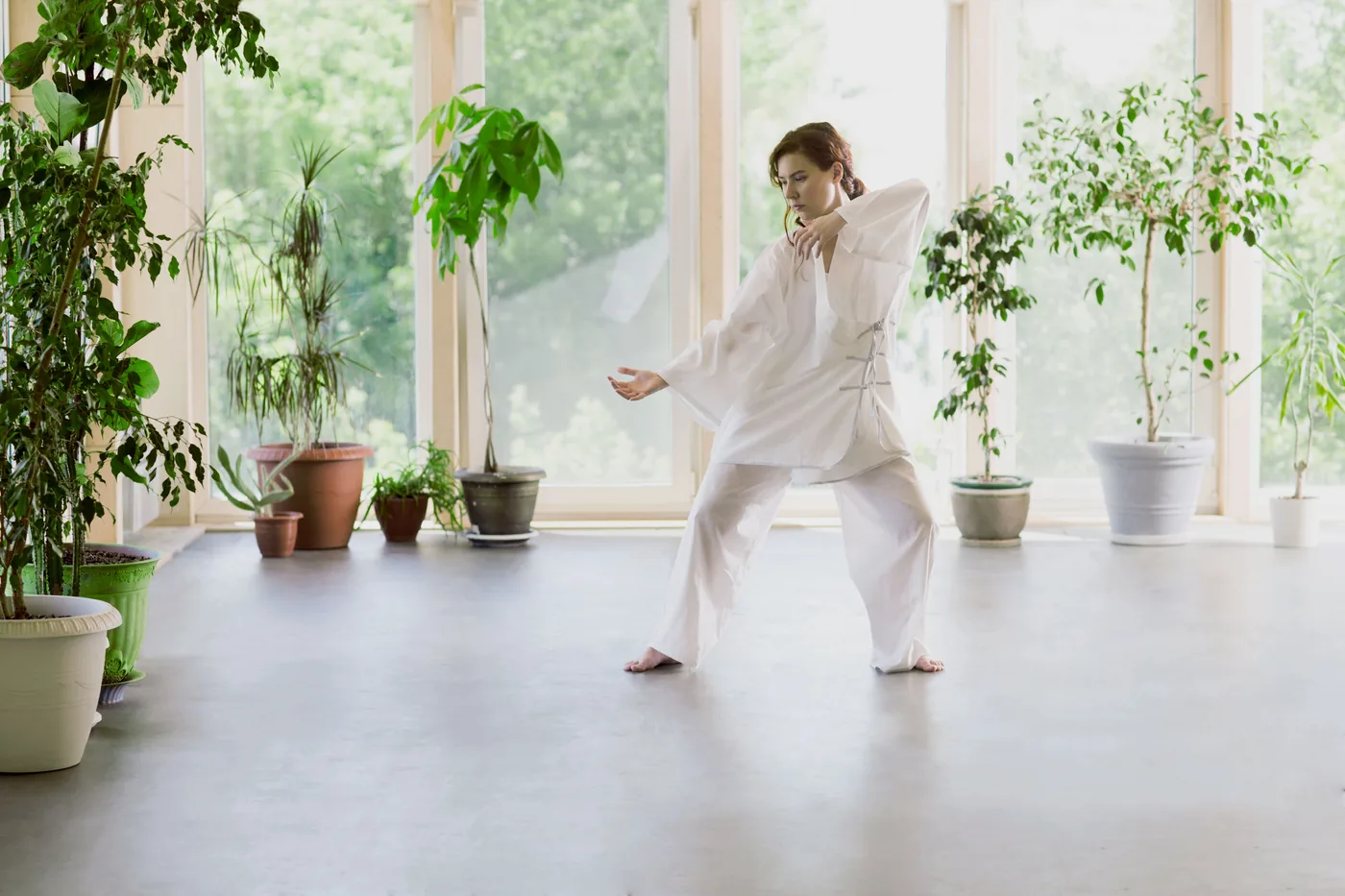 Woman in white tai chi pose in a bright room with potted plants and large windows, looking focused.