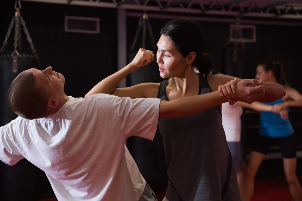 Woman practices martial arts, blocking a man's punch in a gym; others train blurred in the background.
