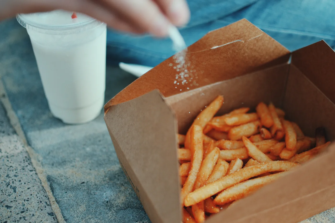 Hand sprinkles salt over fries in a brown takeout box beside a white drink cup on a stone surface.
