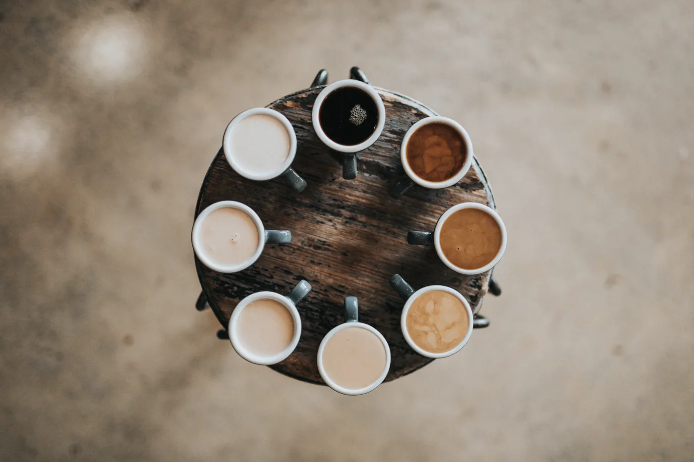 Top-down view of seven coffee mugs in a circle on a rustic round wooden table, with cream, black, and brown drinks.