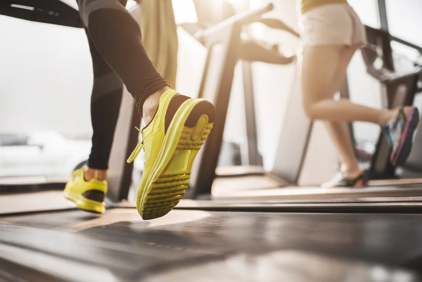Close-up of a runner’s bright yellow shoes on a treadmill, with another person jogging blurred in a sunlit gym