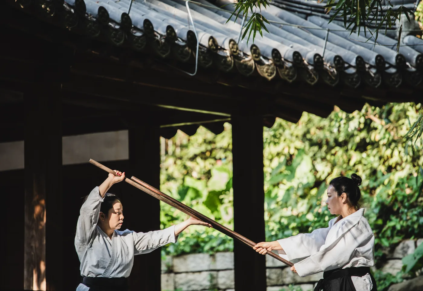 Two women in white martial arts uniforms practice with a long wooden staff under a temple pavilion, greenery behind.