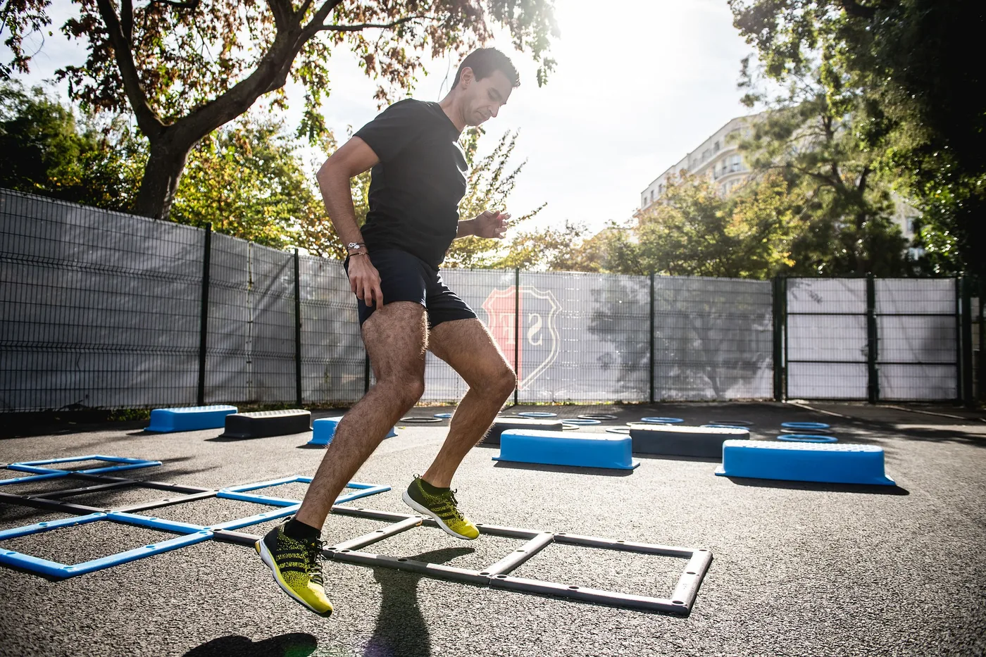Man in black athletic wear jumps through an agility ladder in a sunny outdoor training area with blue hurdles and trees.