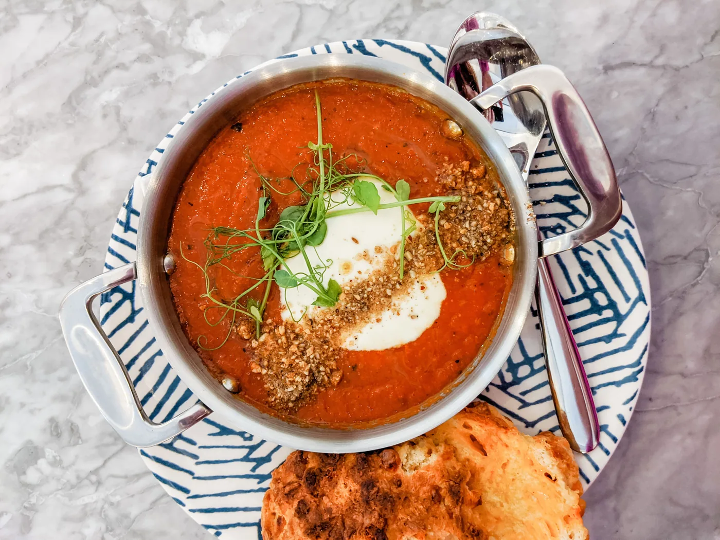Red Lentil soup in a metal bowl with cream and herbs on a blue-patterned plate, served with bread on a marble table