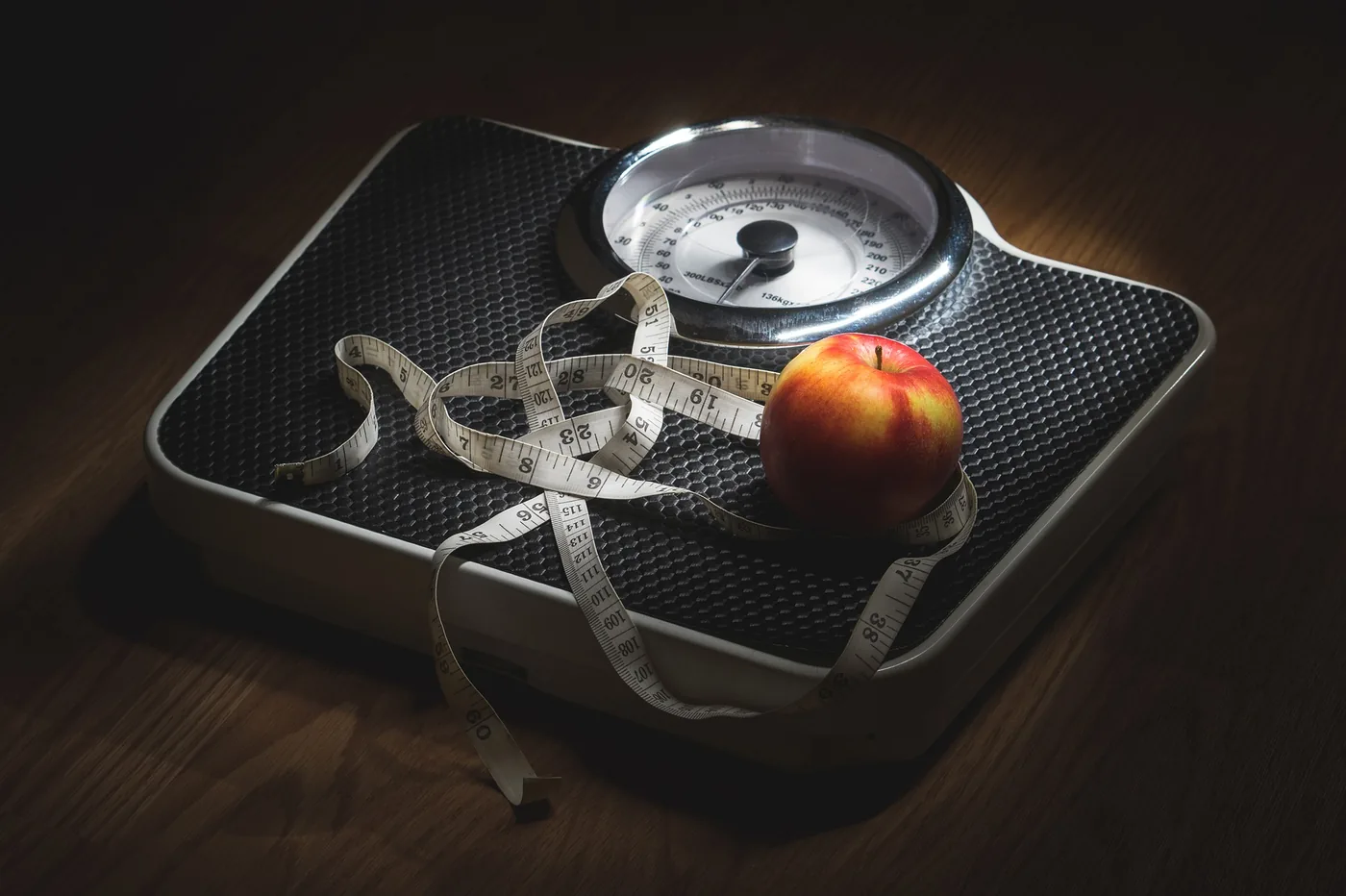 Apple and measuring tape on a bathroom scale, suggesting dieting or weight loss, on a dark wooden floor.