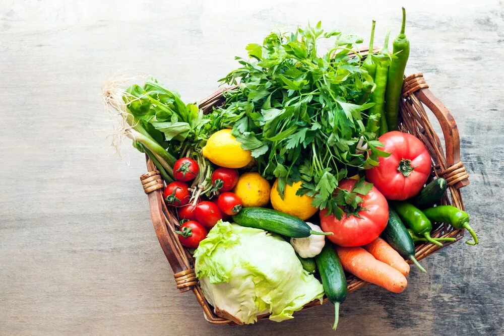 Wicker basket filled with fresh vegetables and herbs—tomatoes, peppers, cucumbers, carrots, lettuce, and lemons on a rustic table.