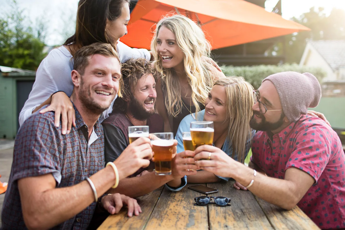 Friends laughing and toasting beers at an outdoor wooden table under an orange patio umbrella, cheerful summer gathering