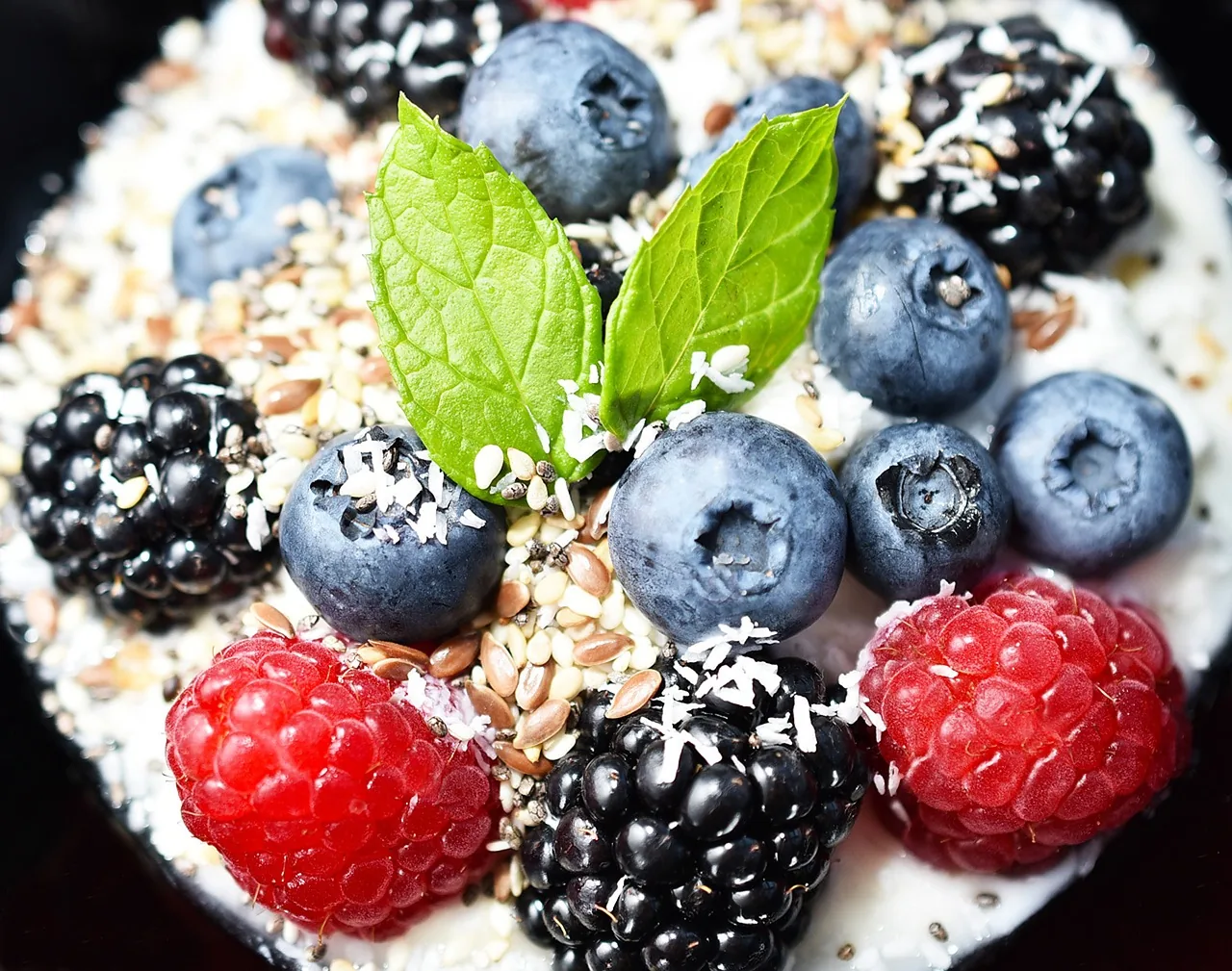 Close-up of yogurt topped with blueberries, blackberries, raspberries, seeds, and fresh mint leaves.