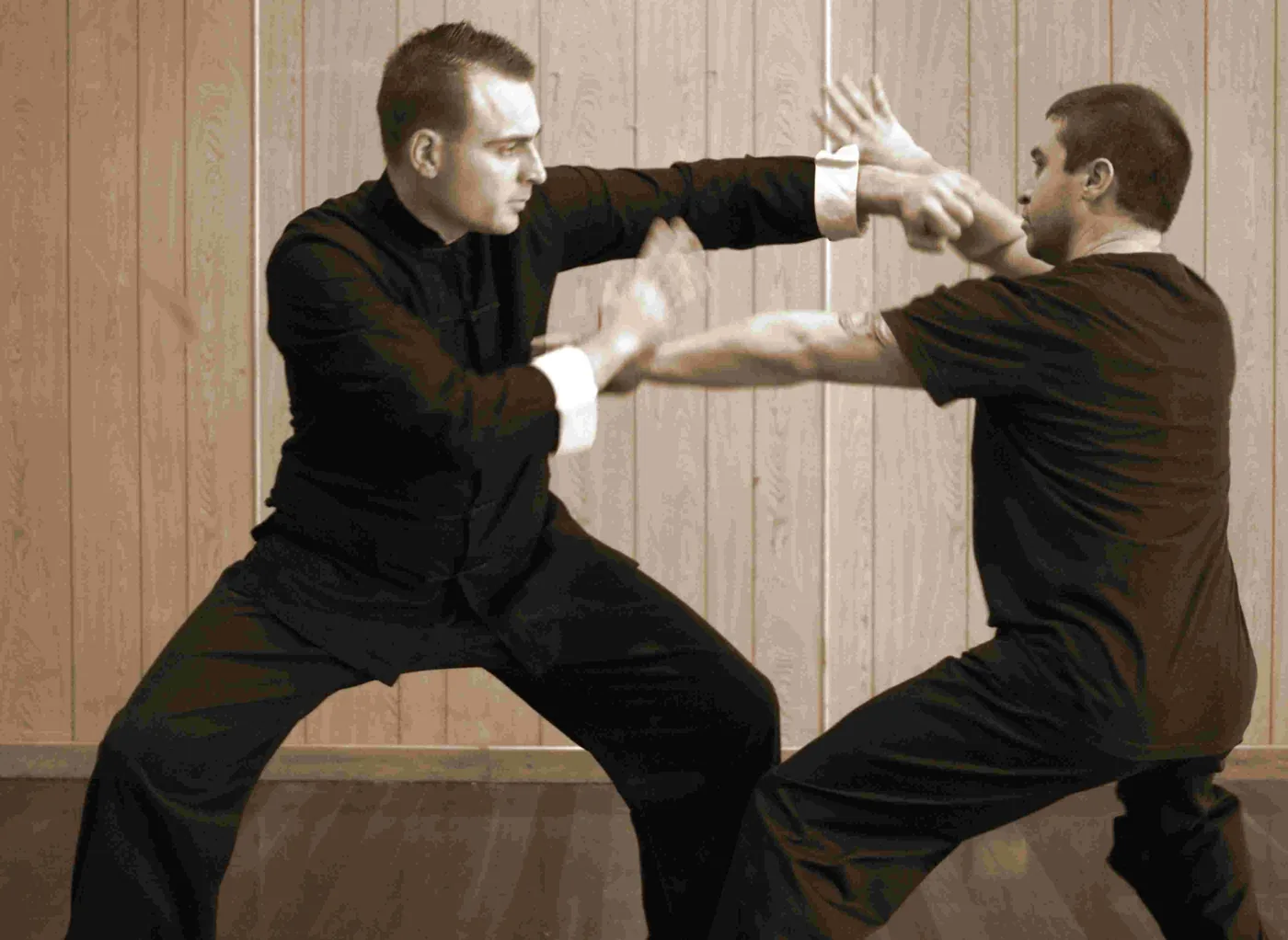 Two men in black martial arts uniforms spar indoors, blocking and striking against a wood-paneled wall.