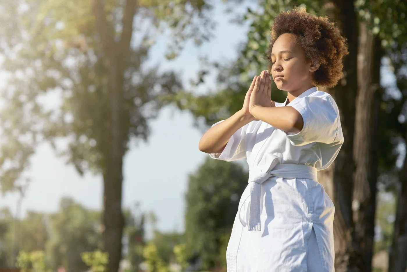 Child in a white karate gi meditates outdoors with eyes closed, hands pressed together, in soft sunlight and trees.
