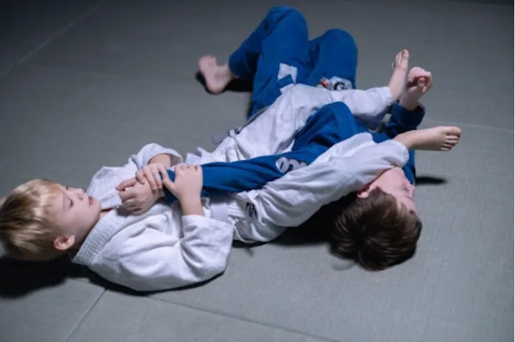 Two children in white and blue martial arts gis grappling on a gray mat during indoor training, focused and playful.