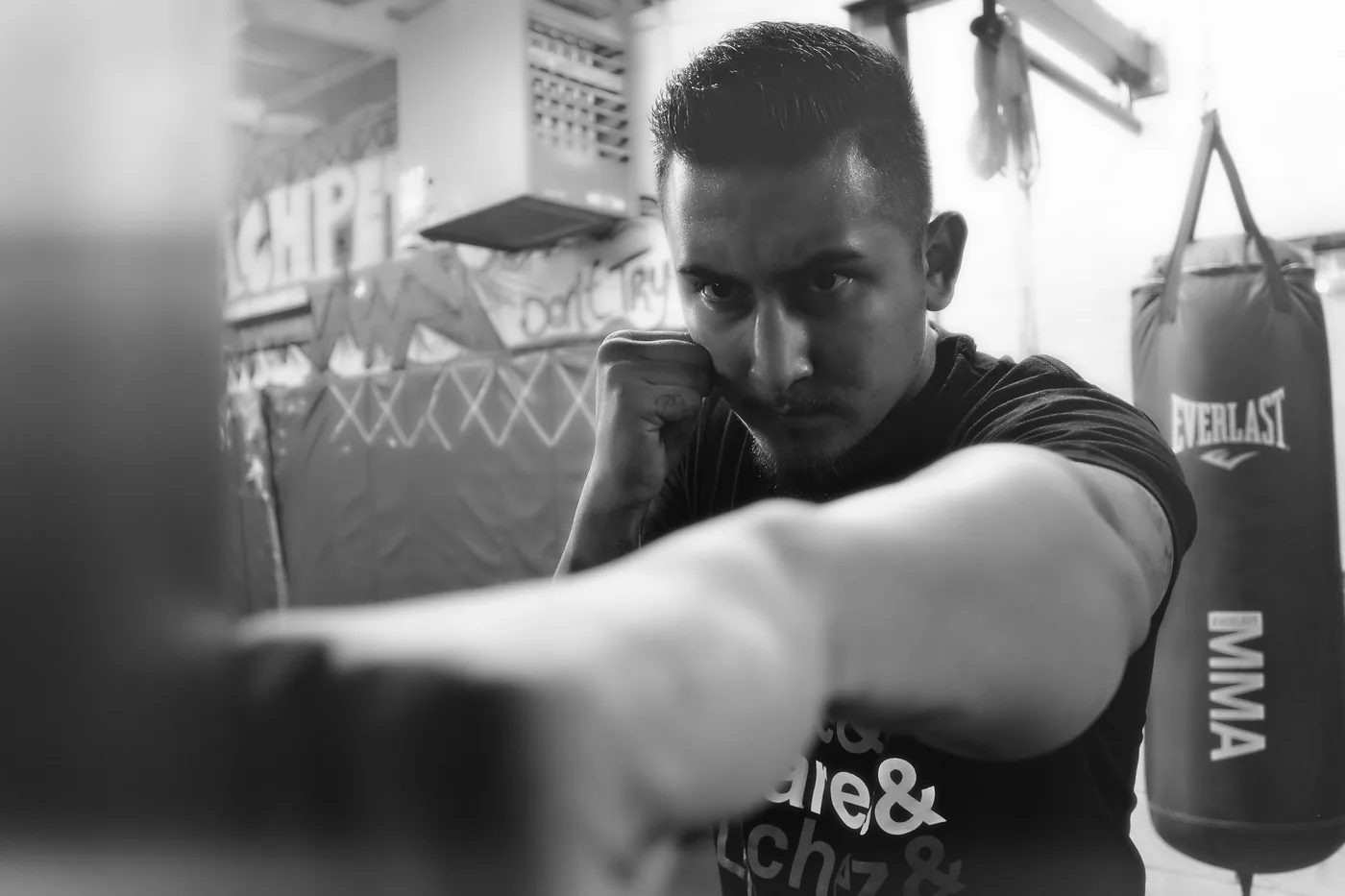 Black-and-white close-up of a focused man throwing a punch in a boxing gym, with an Everlast MMA bag and graffiti behind him.