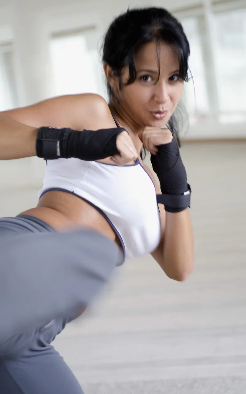 Woman in a martial arts studio throwing a high kick, wearing a white top, gray pants, and black hand wraps.