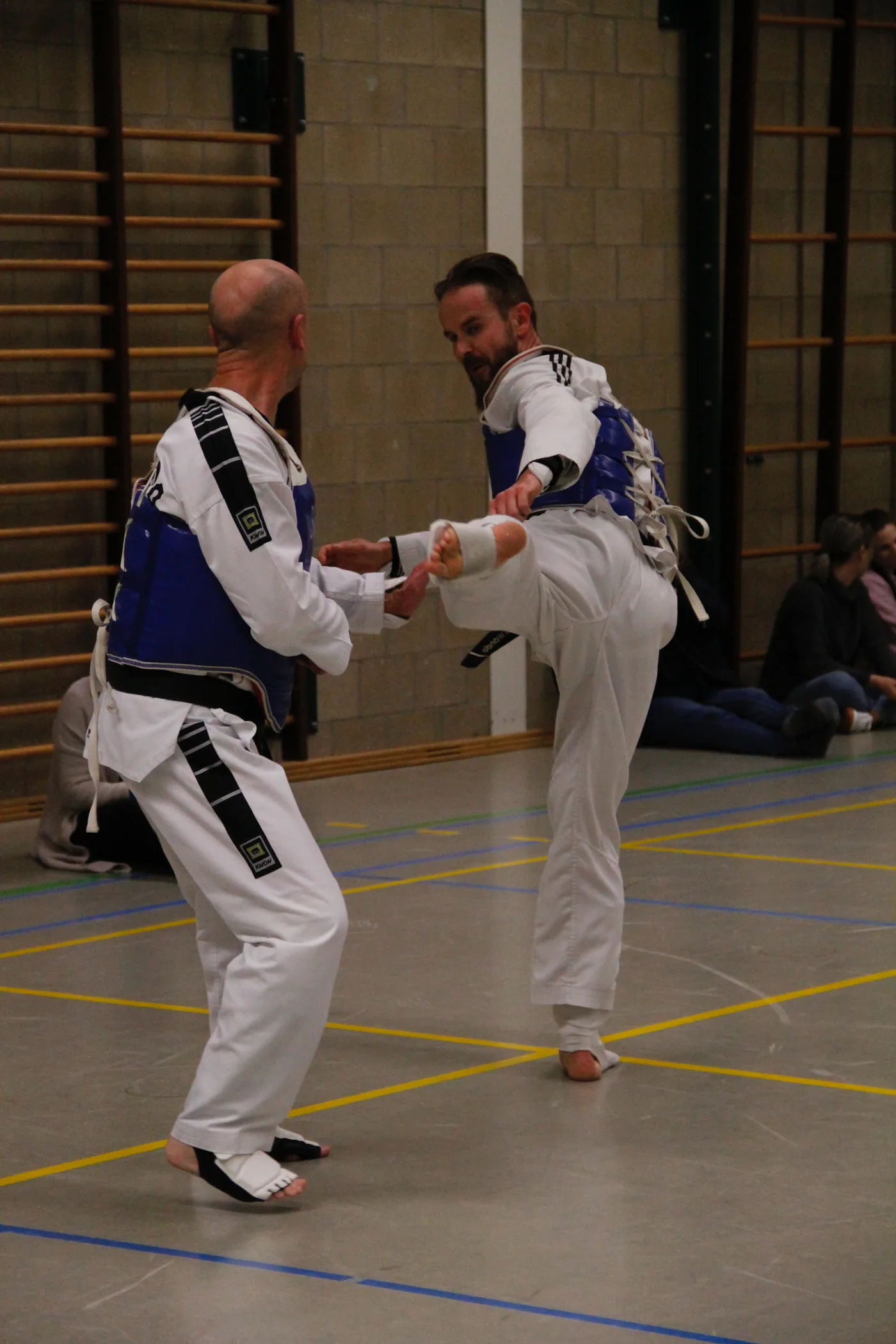 Two taekwondo fighters in white uniforms and blue chest guards spar in a gym, one landing a kick while spectators watch nearby.