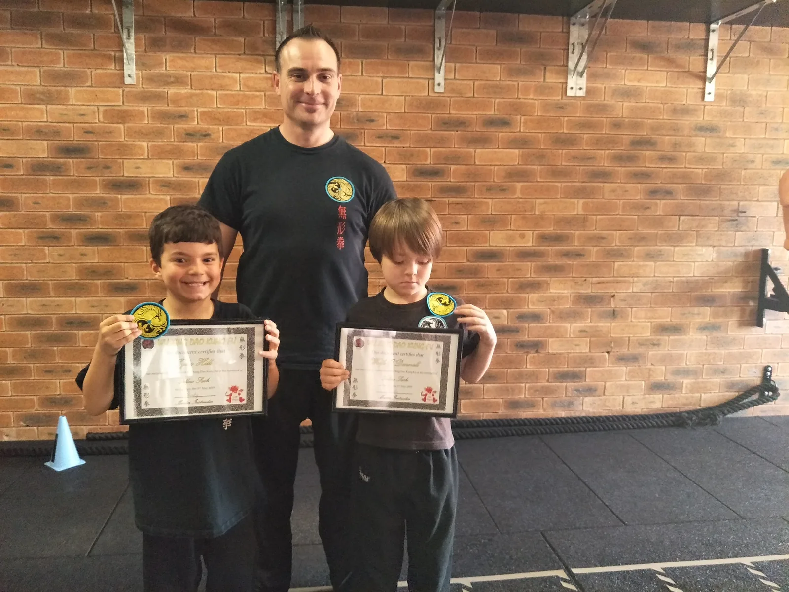 Two young students proudly hold their yellow sash certificates beside their instructor.