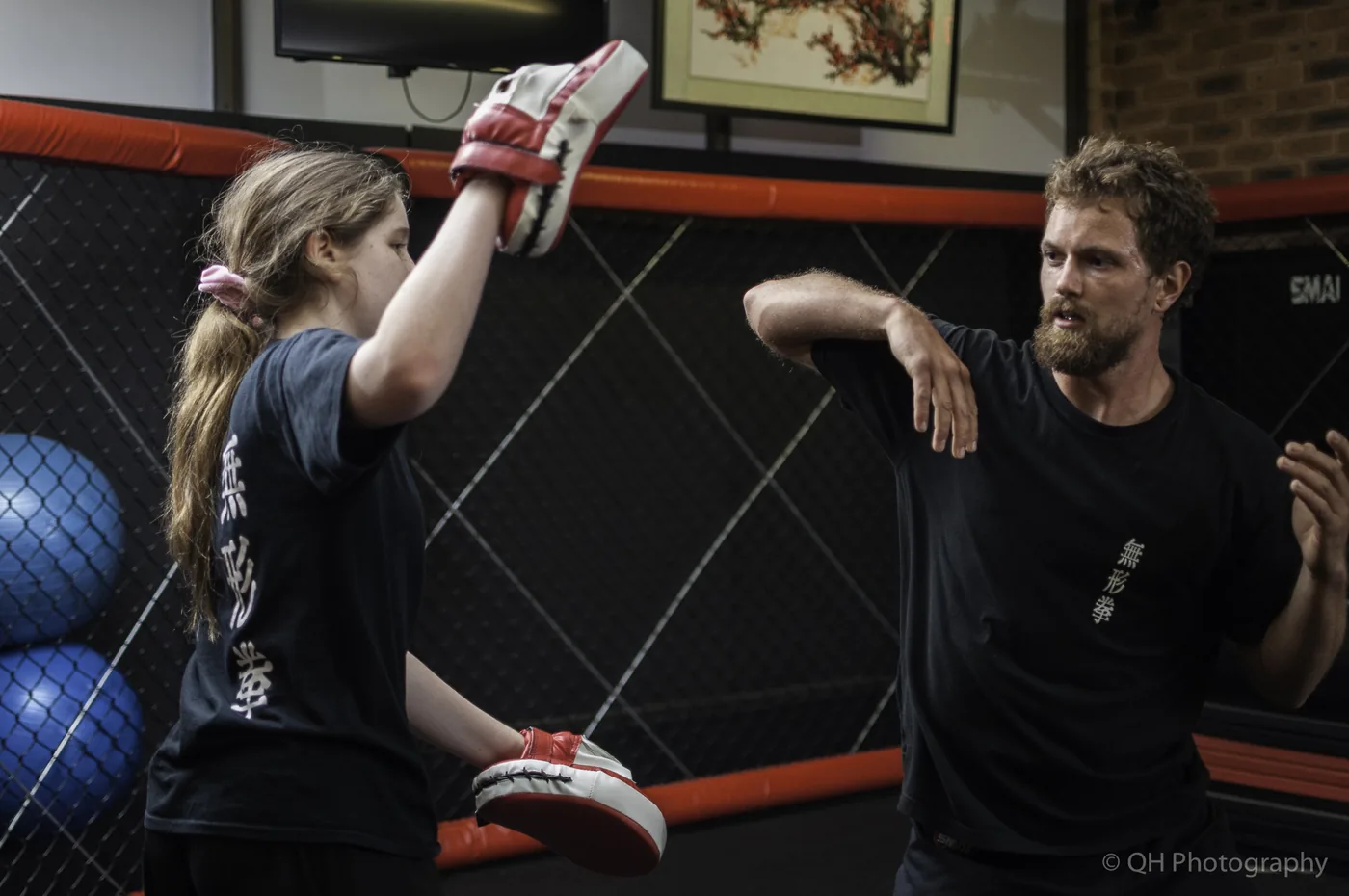 Woman and man practice martial arts in an MMA cage gym, striking red-and-white pads with focused expressions.