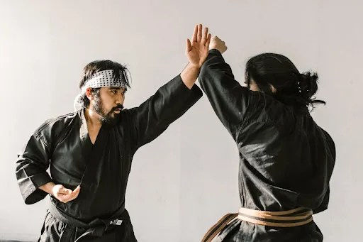 Two martial artists in black gis spar in a bright studio, raising hands to block, focused and intense.