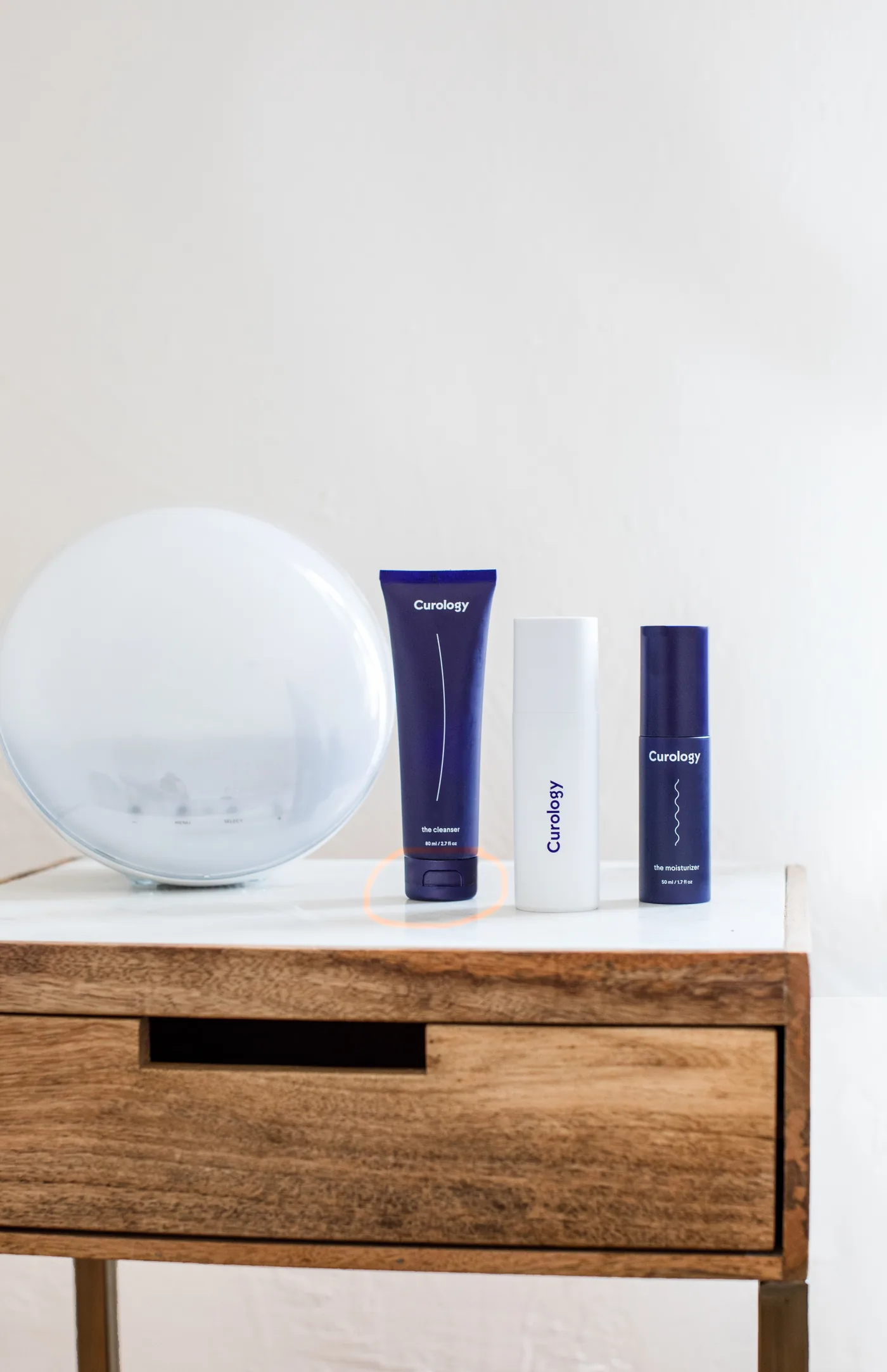 Curology skincare bottles on a wooden table beside a frosted globe lamp, with cleanser and moisturizer text on a bright white background