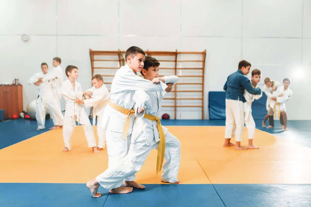 Boys in white judo uniforms practice grappling on orange mats in a bright gym, with wooden wall bars in the background.
