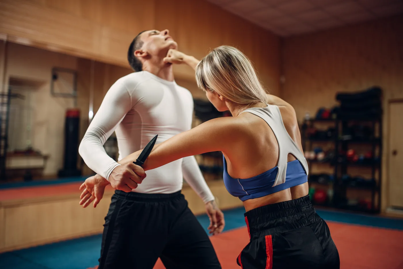 Woman practices knife defense against a man in a martial arts studio, both in athletic wear, focused and tense.