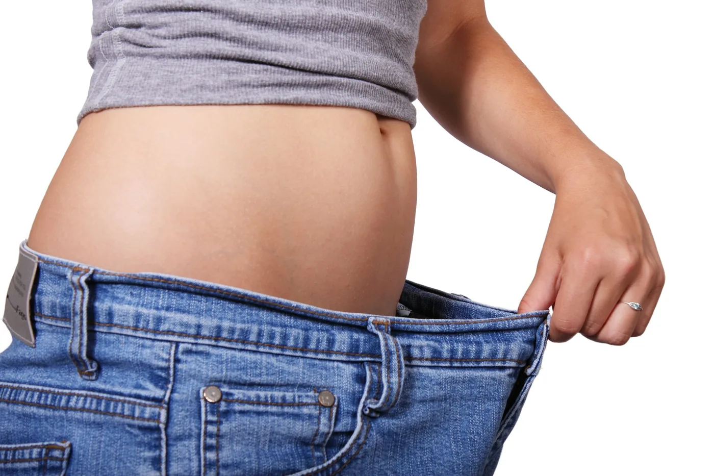 Woman in gray top pulling out oversized blue jeans at the waist on a white background, suggesting weight loss or loose fit