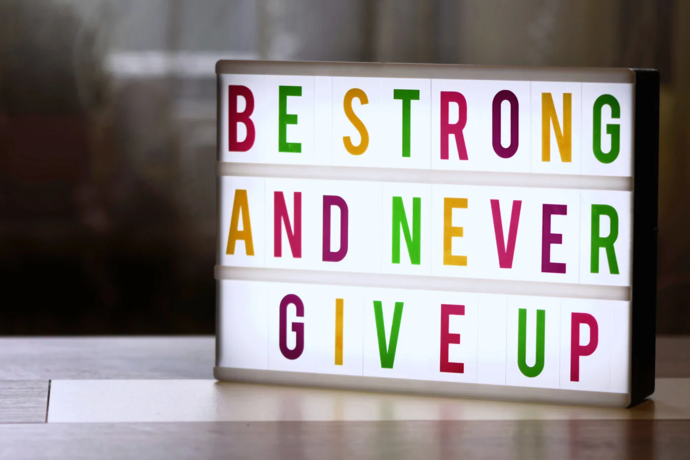 Lightbox sign on a table reads BE STRONG AND NEVER GIVE UP in colorful letters, with a hopeful mood.